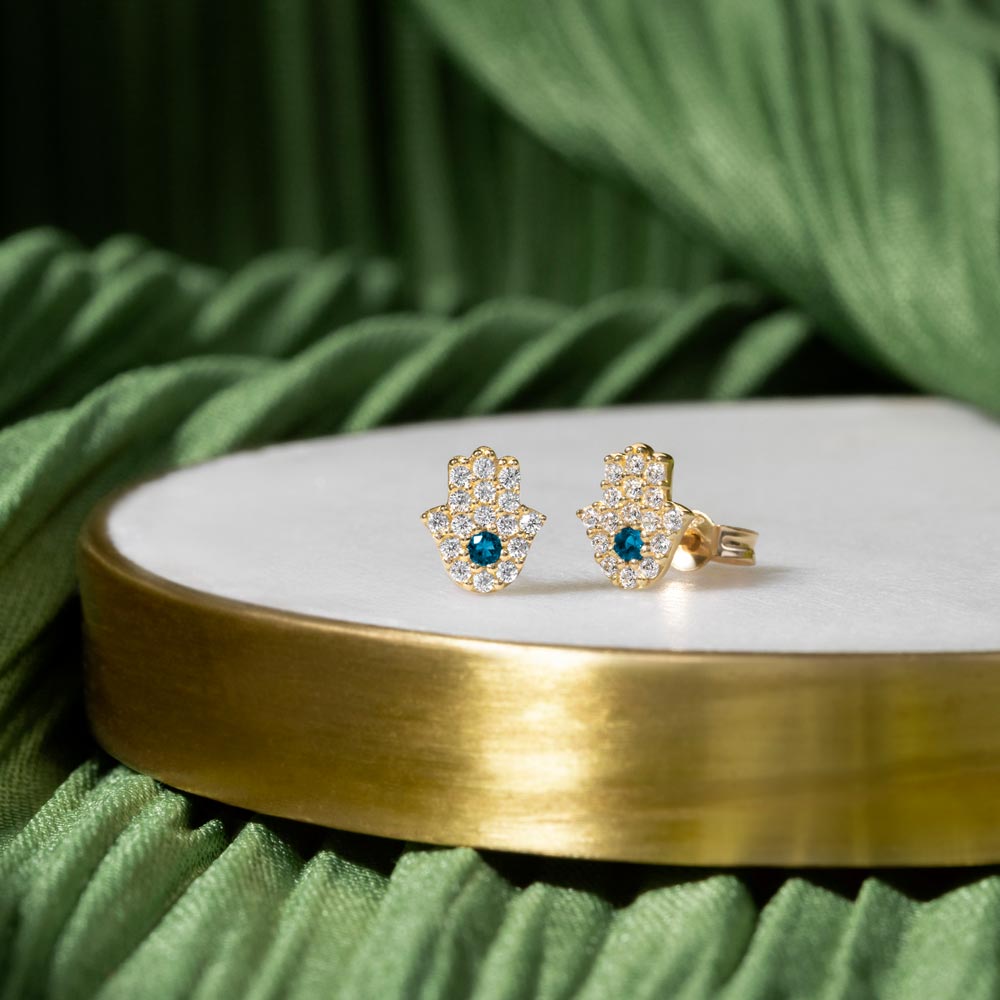 Gold Hamsa hand Studs with cubic zirconia stones placed on a white and green background 