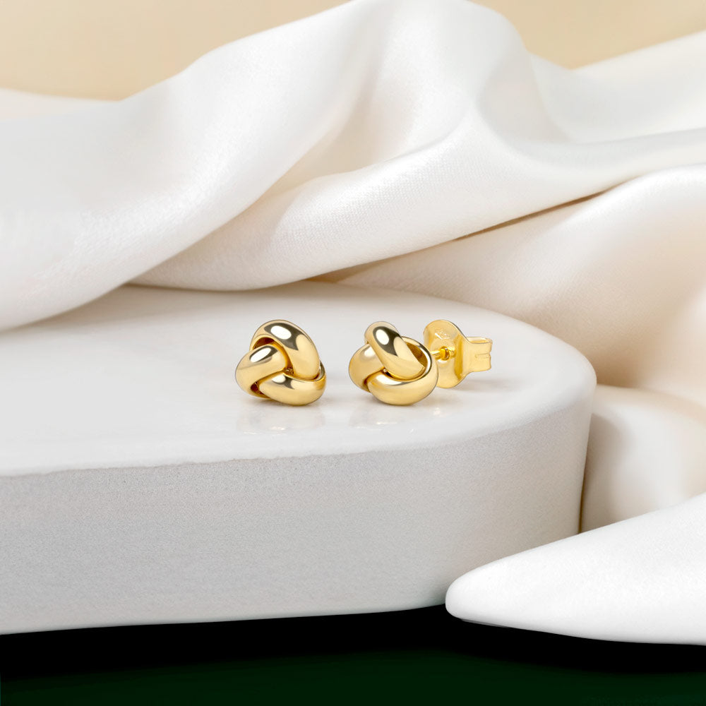 Minimal Gold Knot studs placed on a clean whit tile against cream fabric background