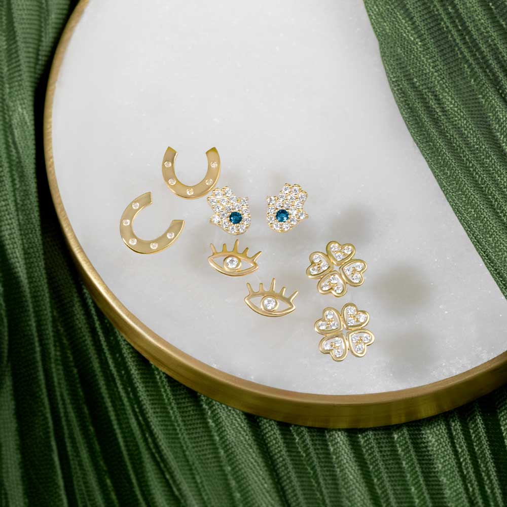 Gold Horseshoe studs, Gold Hamsa hand Studs, Gold evil eye Studs and Gold clover Studs placed on a white and green background 