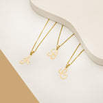 Three gold necklaces displayed on a smooth white surface, each featuring a pendant with a cursive letter from the alphabet:A, B and C