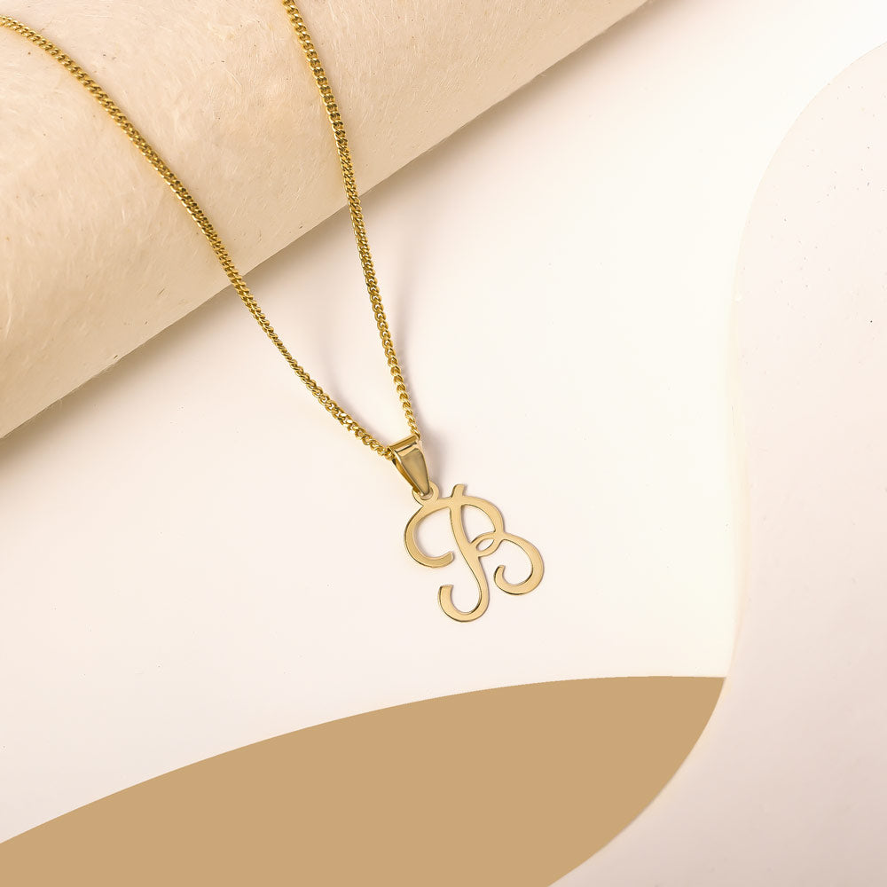 A delicate gold necklace is pictured with an alphabet pendant in the form of the letter 'B' resting on a cream-colored surface