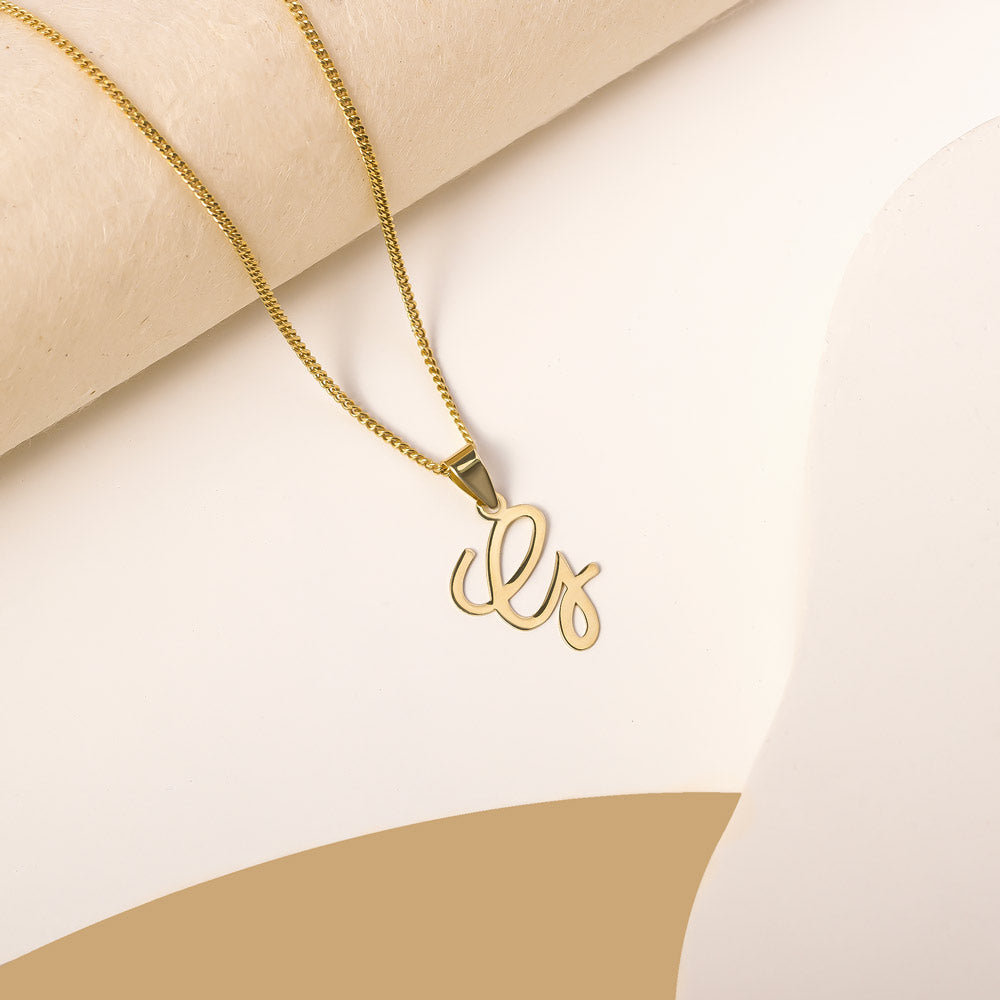 A delicate gold necklace is pictured with an alphabet pendant in the form of the letter 'G' resting on a cream-colored surface