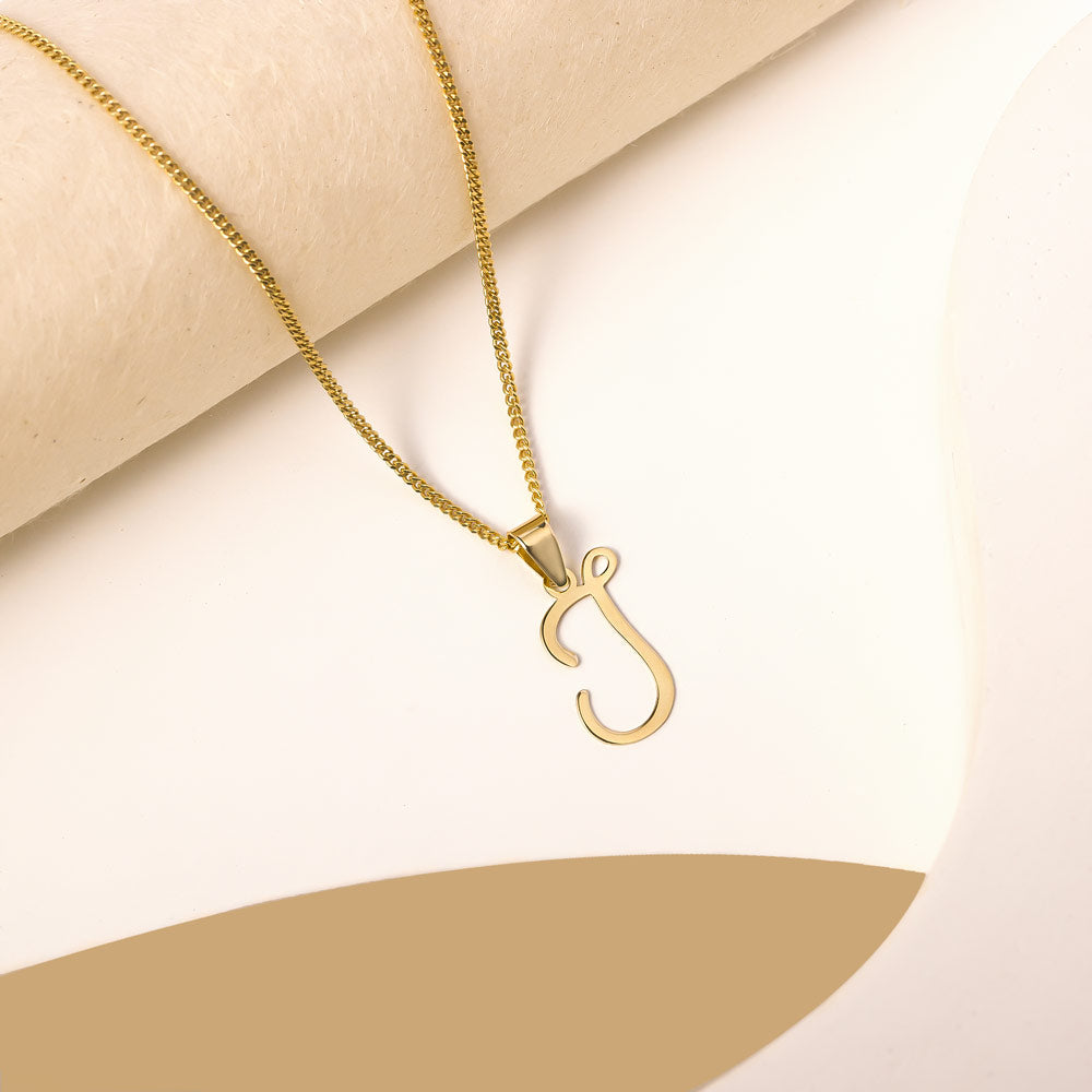 A delicate gold necklace is pictured with an alphabet pendant in the form of the letter 'J' resting on a cream-colored surface