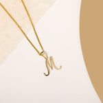 A delicate gold necklace is pictured with an alphabet pendant in the form of the letter 'M' resting on a cream-colored surface