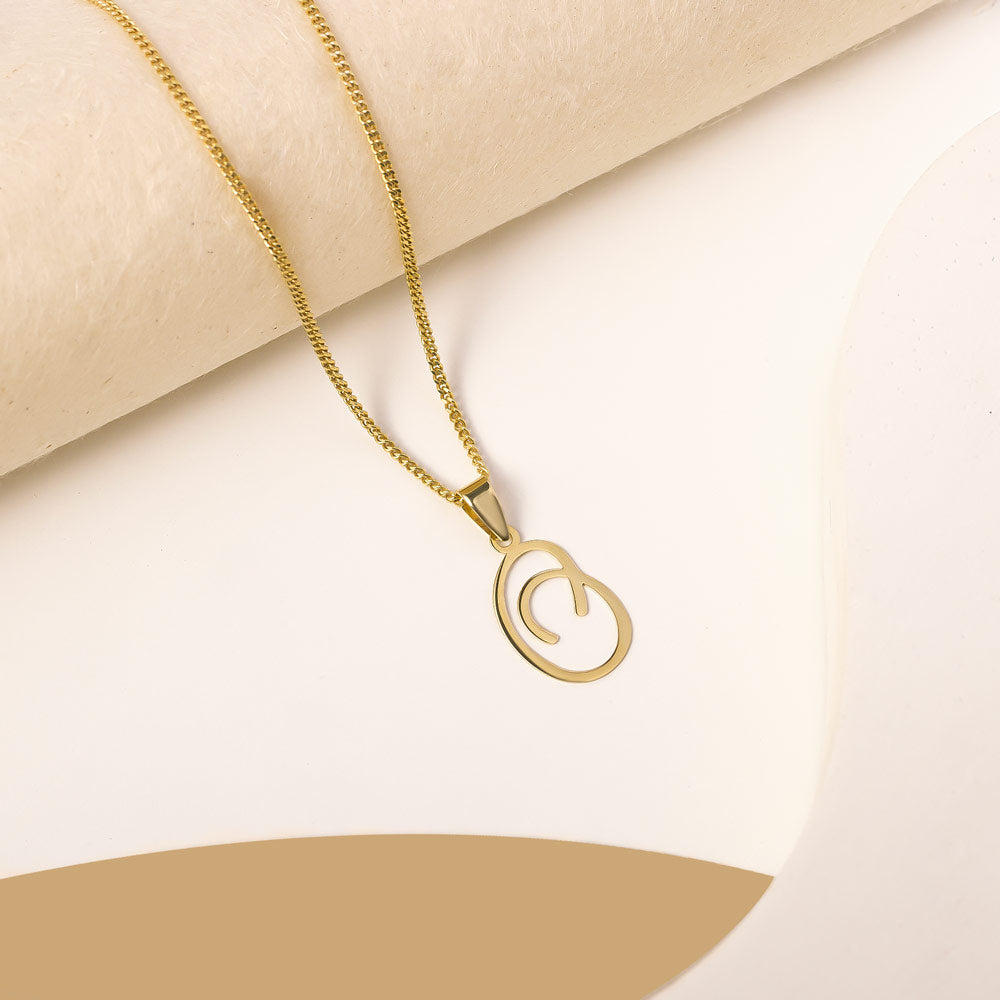 A delicate gold necklace is pictured with an alphabet pendant in the form of the letter 'O' resting on a cream-colored surface