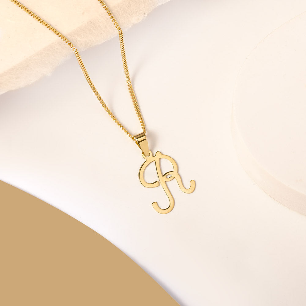 A delicate gold necklace is pictured with an alphabet pendant in the form of the letter 'R' resting on a cream-colored surface