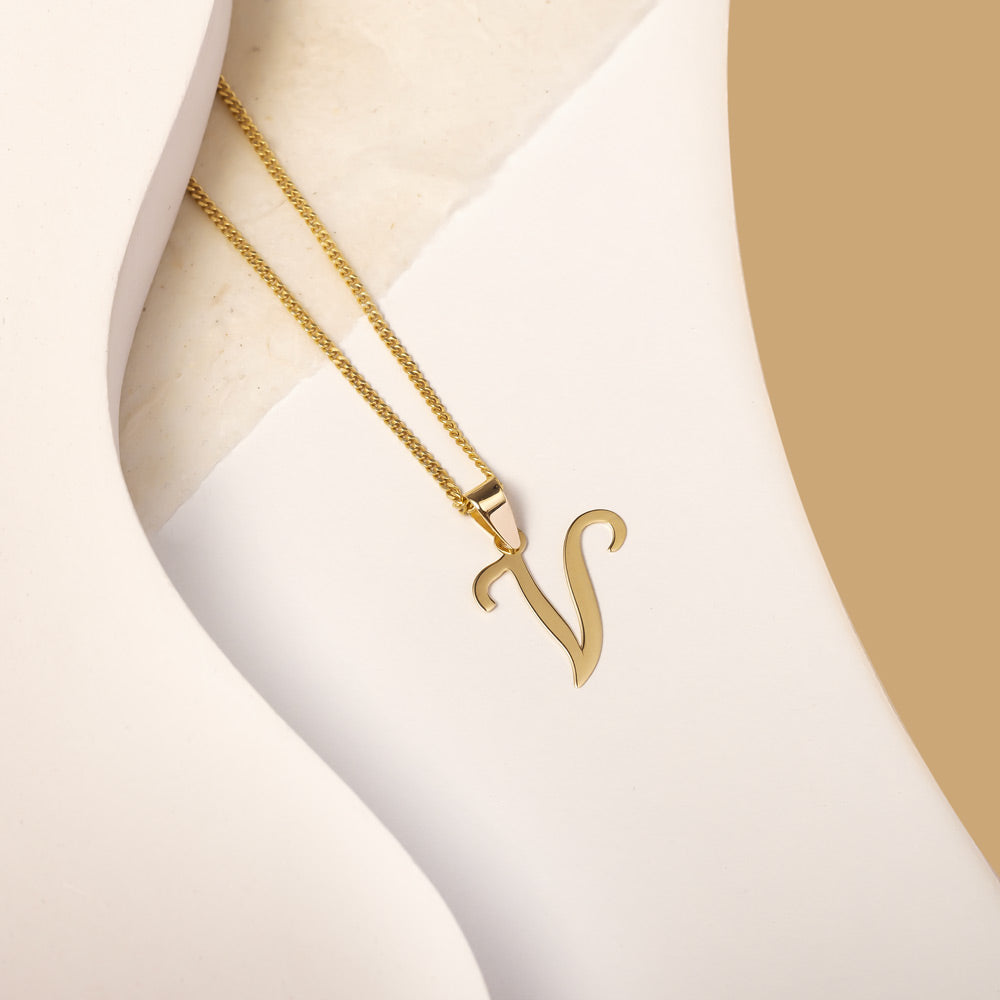 A delicate gold necklace is pictured with an alphabet pendant in the form of the letter 'V' resting on a cream-colored surface