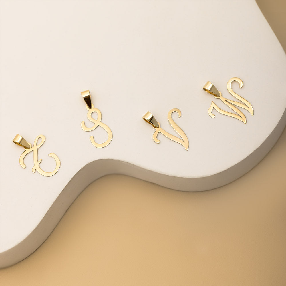 Four gold pendants in the shapes of the alphabet letters "T," "S," "V," and "W" are arranged on a white, wavy surface against a beige background