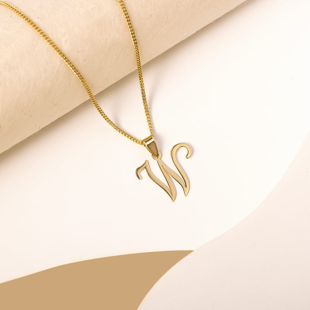 A delicate gold necklace is pictured with an alphabet pendant in the form of the letter 'W' resting on a cream-colored surface