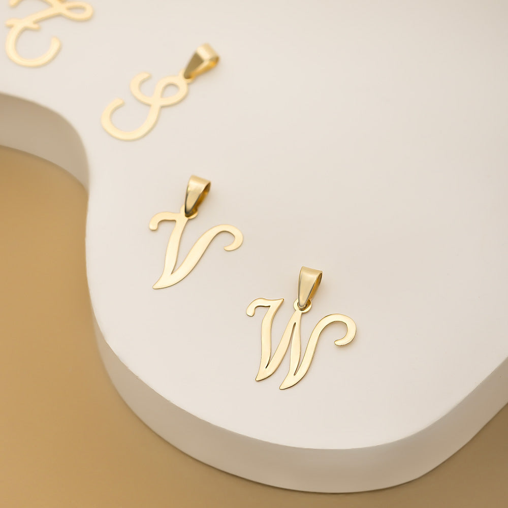 Three gold alphabet letter pendants are displayed on a white and beige surface featuring shiny gold alphabets S, V and W