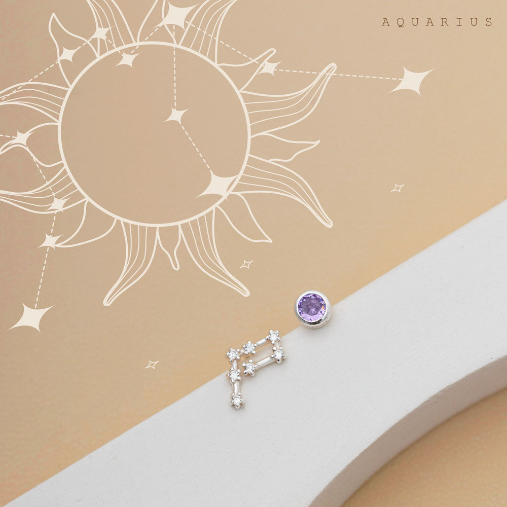 A pair of  birthstone constellation studs sits on a beige background  with sun and constellation motifs. One earring resembles the Aquarius constellation, while the other showcases a single round purple gemstone