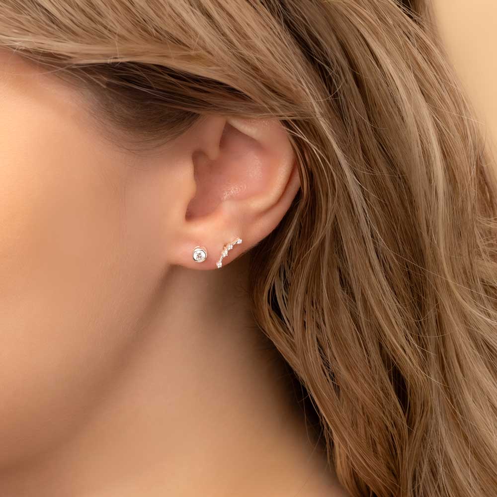 Close-up of a woman's ear wearing two earrings: a small stud earring featuring a diamond birthstone and a stunning Aries constellation-shaped piece with clear stones