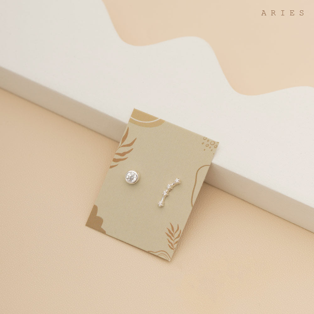 A small card rests on a beige surface, On the card are two delicate earrings: one stud with a birthstone gem and another with constellation shape. The word "ARIES" is placed in the top right corner.