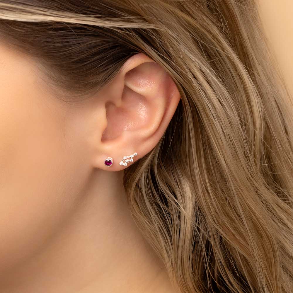 Close-up of a woman's ear wearing two earrings: a small stud earring featuring a ruby birthstone and a stunning Cancer constellation-shaped piece with clear stones