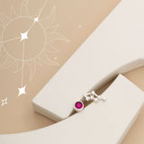 A single earring with a small red gemstone and a Cancer constellation design rests on a curved, white geometric surface. A faint, stylized sun and stars are illustrated on the beige background.