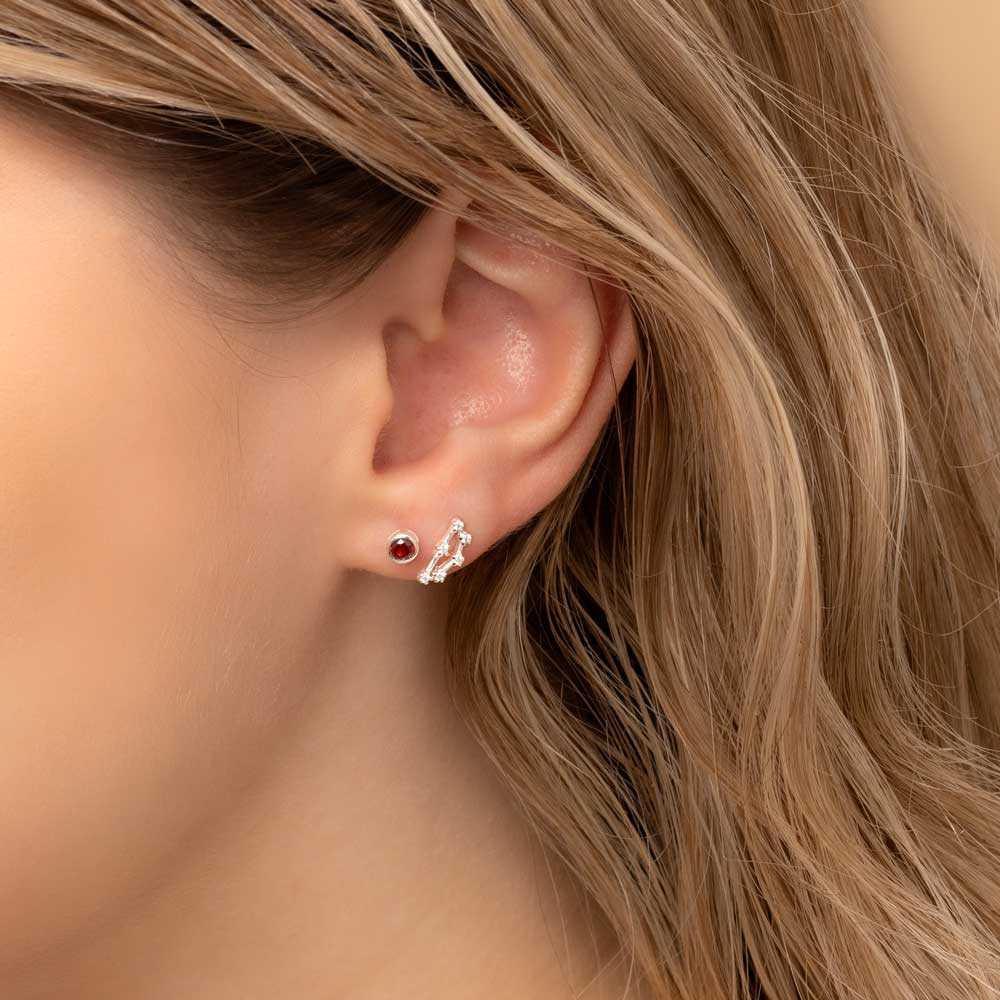 Close-up of a woman's ear wearing two earrings: a small stud earring featuring a garnet birthstone and a stunning Capricorn constellation-shaped piece with clear stones