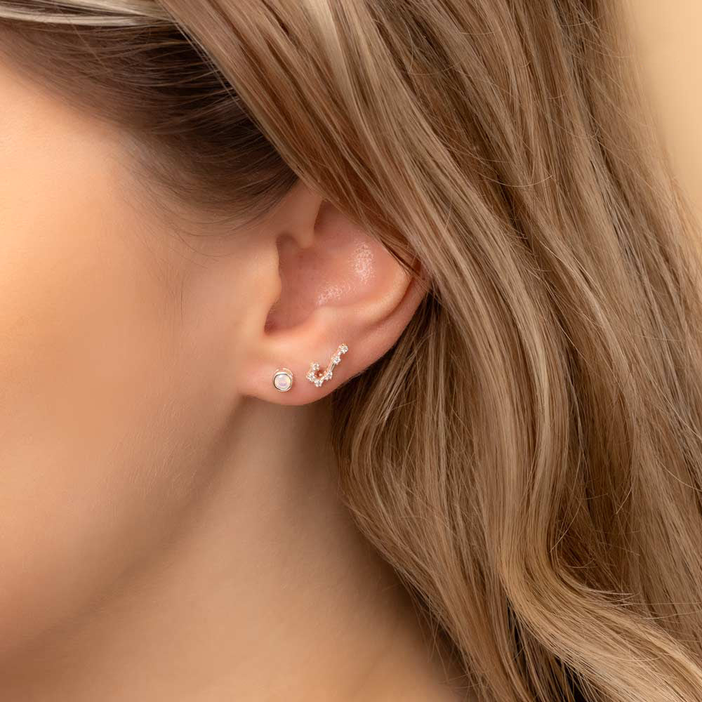 Close-up of a woman's ear wearing two earrings: a small stud earring featuring a moonstone birthstone and a stunning Gemini constellation-shaped piece with clear stones
