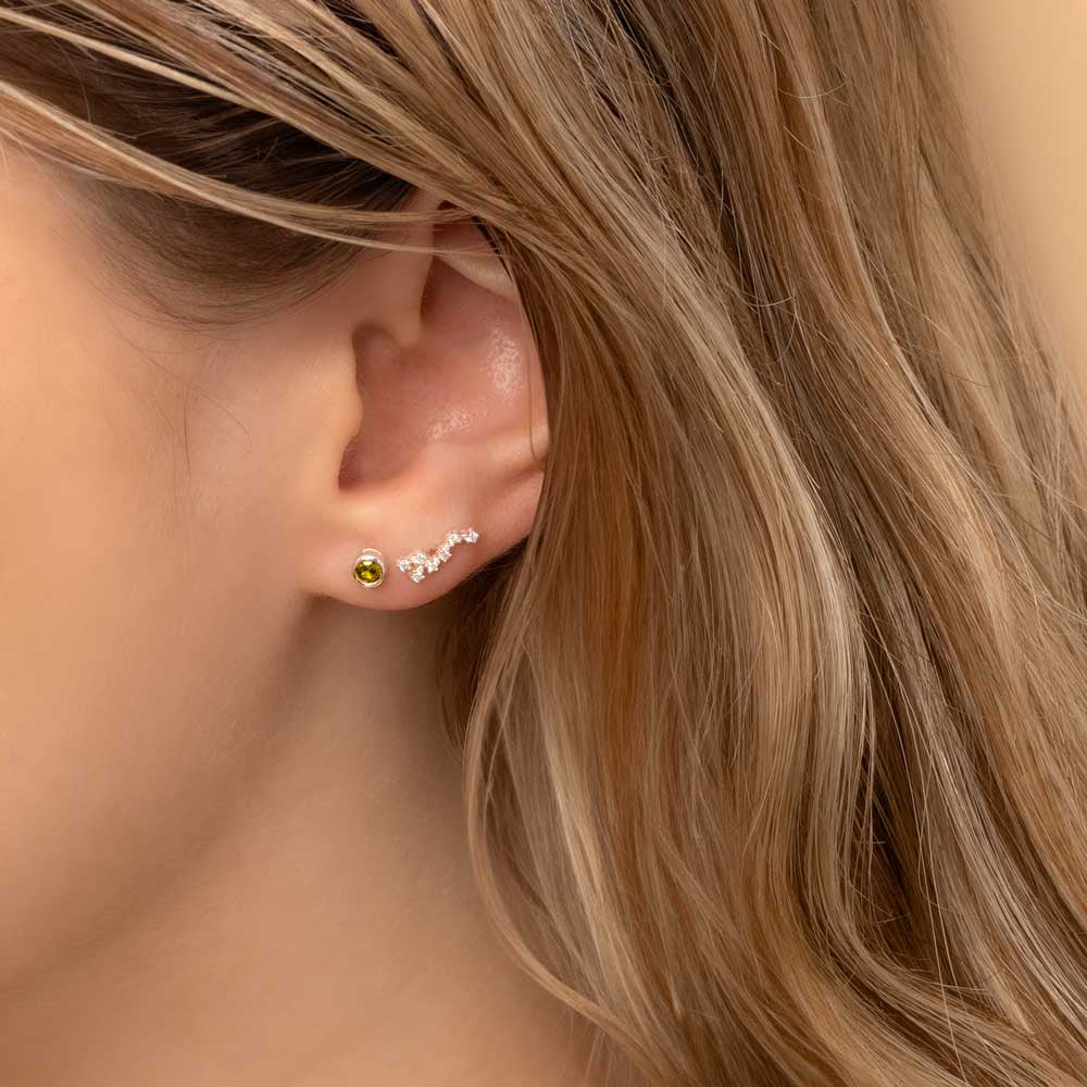 Close-up of a woman's ear wearing two earrings: a small stud earring featuring a peridot birthstone and Leo constellation-shaped piece with clear stones
