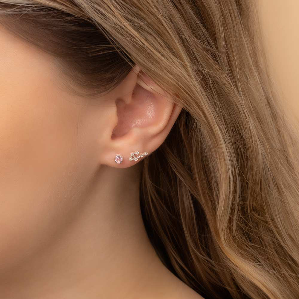 Close-up of a woman's ear wearing two earrings: a small stud earring featuring a pink tourmaline birthstone and a stunning Libra constellation-shaped piece with clear stones