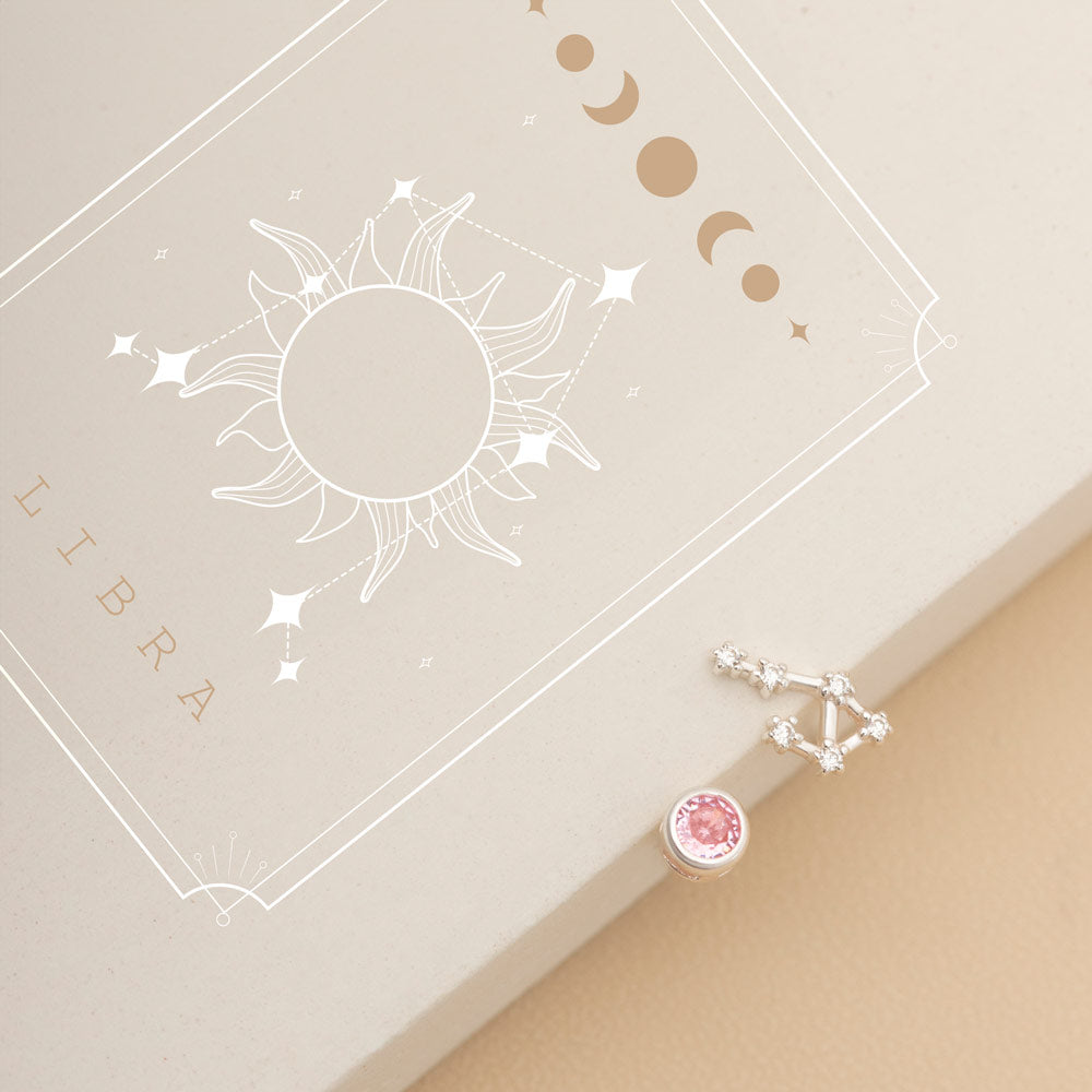  A pair of mismatched studs, One earring showcases the Libra birthstone, a pink gemstone in a silver setting, while the other features constellation-like studs made of small, clear stones.