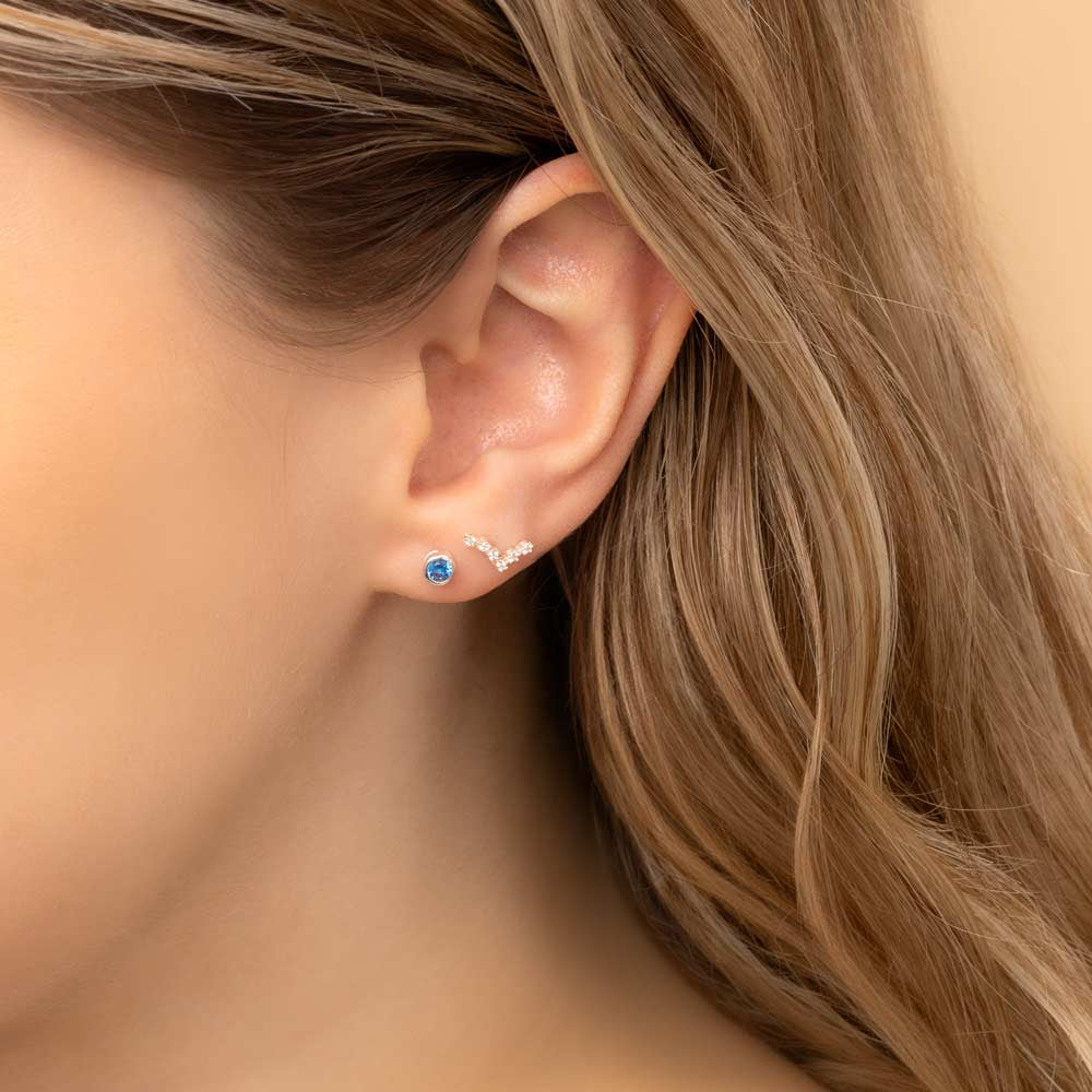 Close-up of a woman's ear wearing two earrings: a small stud earring featuring a aquamarine birthstone and a stunning Pisces constellation-shaped piece with clear stones