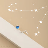 A minimalistic image features Pisces birthstone studs adorned with a blue gem and small crystals. Above them, white star illustrations form the Pisces constellation on a beige background.