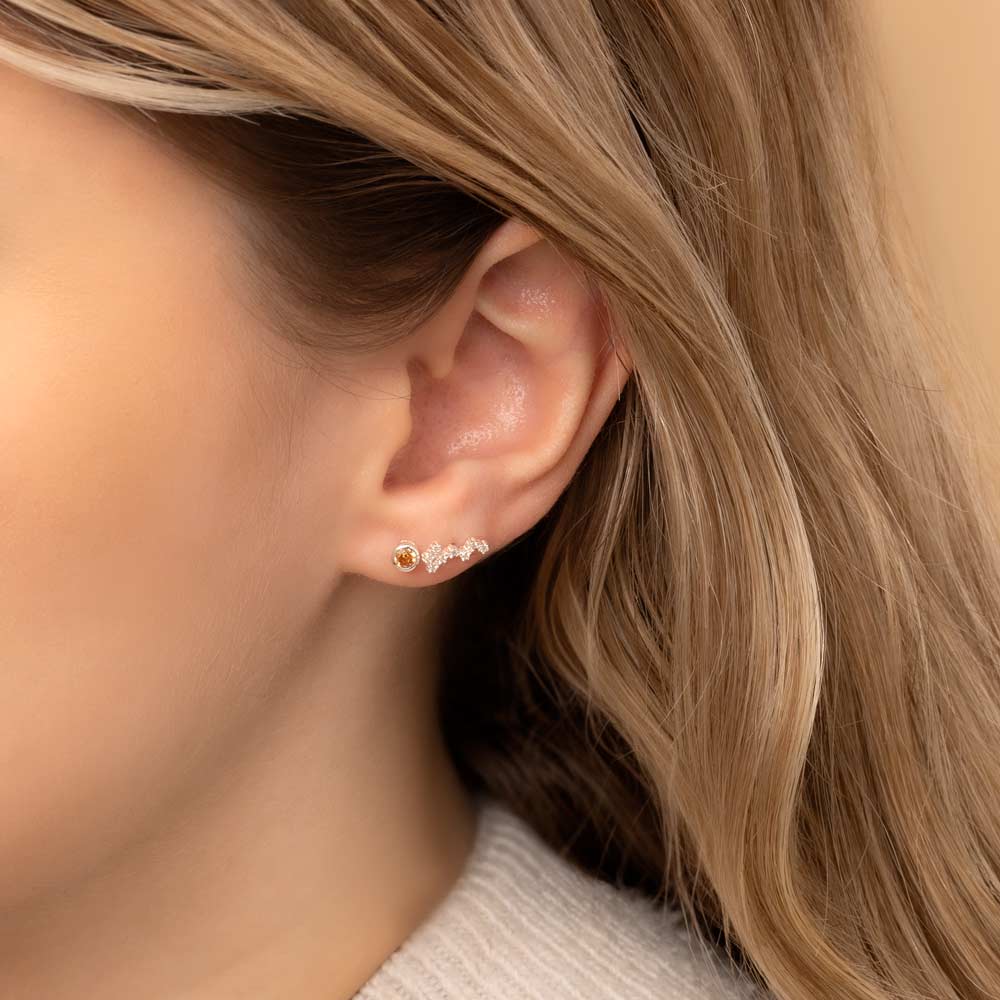 Close-up of a woman's ear wearing two earrings: a small stud earring featuring a topaz birthstone and a stunning Scorpio constellation-shaped piece with clear stones