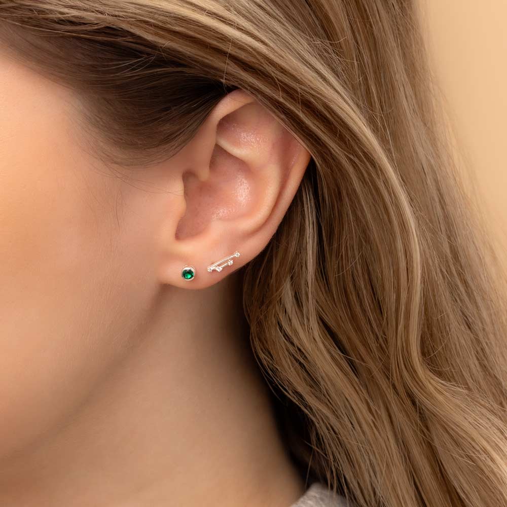 Close-up of a woman's ear wearing two earrings: a small stud earring featuring a emerald birthstone and a stunning Taurus constellation-shaped piece with clear stones