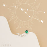 Minimalist image featuring a pair of earrings on a beige surface with a Taurus constellation graphic in the background. One earring is a  constellation and the other showcases the Taurus birthstone with a single green gem