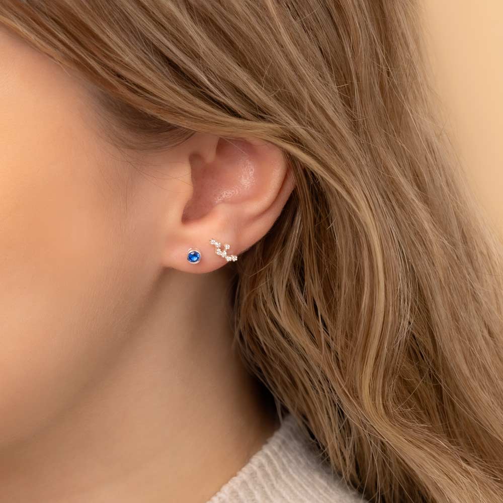 Close-up of a woman's ear wearing two earrings: a small stud earring featuring a sapphire birthstone and a stunning Virgo constellation-shaped piece with clear stones